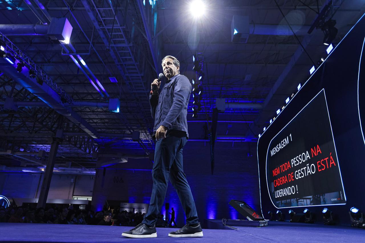 Vista ampla do palco com Adriano Lima palestrando e o telão exibindo “Nem toda pessoa na cadeira de gestão está liderando!”