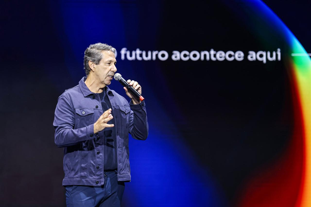 Adriano Lima palestrando no palco, de microfone na mão, diante de um telão azul com a frase “o futuro acontece aqui”